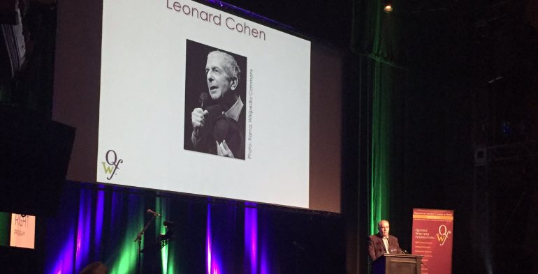 Older man speaking in front of projection of Leonard Cohen's photo
