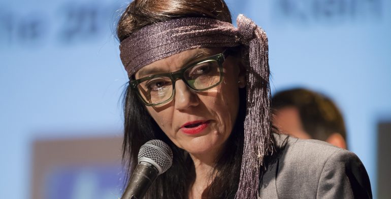 Woman wearing a purple scarf around her head reading into a microphone