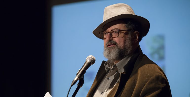 Middle aged man wearing a fedora reading at a microphone